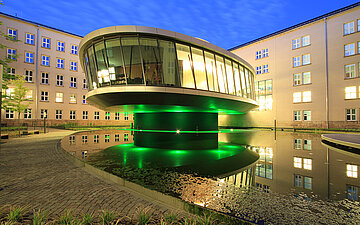 Foto: Bundessozialgericht in Kassel: Modernes, rundes Glasgeb&auml;ude mit gr&uuml;ner Beleuchtung, das &uuml;ber einem Wasserbecken schwebt, nachts in einem Innenhof zwischen hellen Verwaltungsgeb&auml;uden.