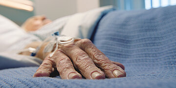 Das Foto zeigt eine alte Hand in Nahaufnahme auf einer blauen Krankenhaus-Bettdecke