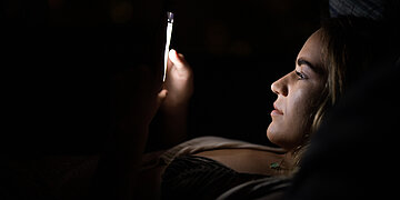 Foto: Frau liegt im Dunkeln und schaut auf den beleuchteten Bildschirm eines Smartphones.