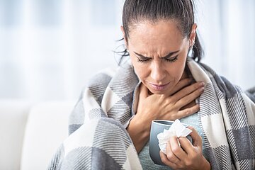 Eine Frau sitzt in eine Decke geh&uuml;llt mit einer Tasse Tee auf der Couch und hat augenscheinlich Halsschmerzen.