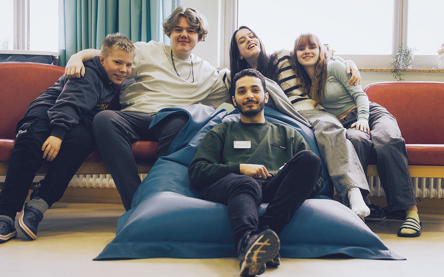 Foto des Pflegers Khaled, wie er in einem Sitzsatz vor zwei fröhlichen schauenden Jungen und Mädchen sitzt, die auf einer Couch Platz genommen haben.  