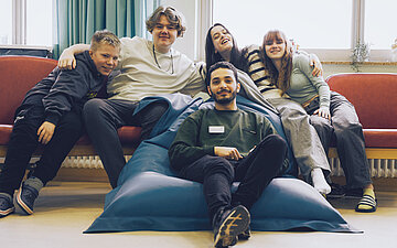Foto des Pflegers Khaled, wie er in einem Sitzsatz vor zwei fr&ouml;hlichen schauenden Jungen und M&auml;dchen sitzt, die auf einer Couch Platz genommen haben. 