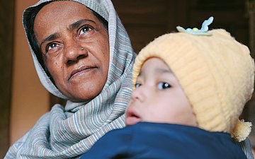 Eine Frau mit Kopftuch und Nasenpiercing hält ein Baby im Arm. Das Baby hat eine blaue Jacke an und eine gelbe Mütze auf.