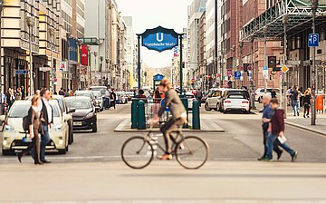 Eine Berliner Straße am U-Bahnhof "Französische Straße"; Personen überqueren zu Fuß oder zu Fahrrad die befahrene Straße.