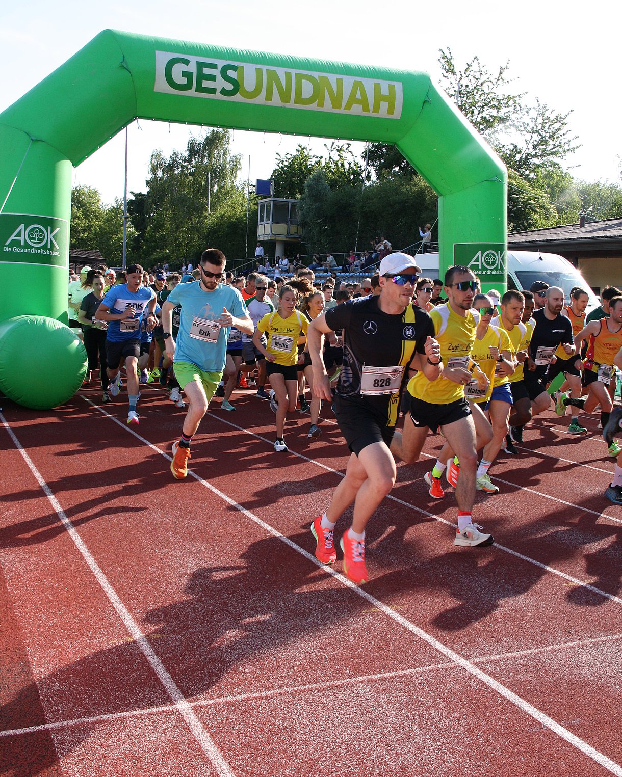 Einige Läufer sind beim Start zum 2. AOK Firmenlauf Bühl zu sehen. Sie laufen dabei auf einer Tartanbahn  durch ein grünes Tor.