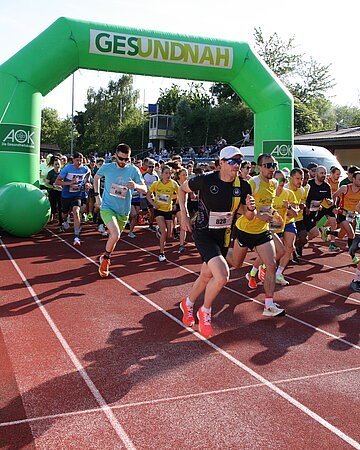 Einige L&auml;ufer sind beim Start zum 2. AOK Firmenlauf B&uuml;hl zu sehen. Sie laufen dabei auf einer Tartanbahn durch ein gr&uuml;nes Tor.