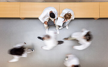 Foto: Bild von oben auf Menschen im wei&szlig;en Kittel, einige bewegen sich, zwei stehen, mit Tablet und Block, nebeneinander und beraten sich.
