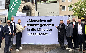 Eine Gruppe steht vor einem gro&szlig;en Banner, auf dem steht: "Menschen mit Demenz geh&ouml;ren in die Mitte der Gesellschaft."