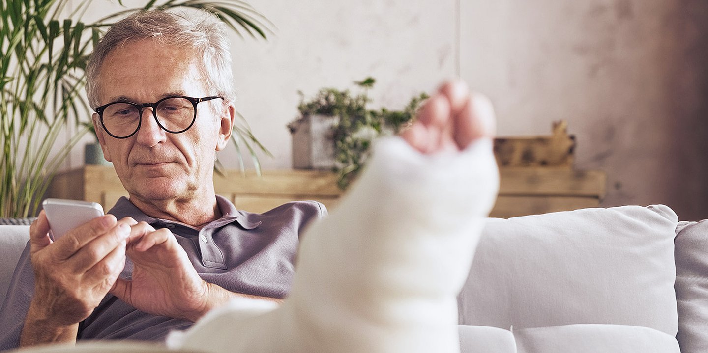 Ein Mann mit einem bandagierten, hochgelegten Fuß sitzt auf dem Sofa und schaut auf sein Handy.