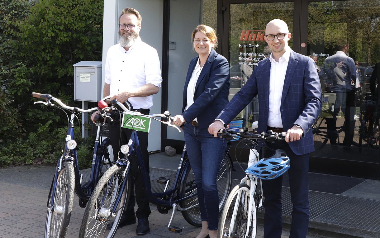 Foto zeigt drei Personen, die jeweils mit dem Fahrrad nebeneinander vor einem Gebäude stehen und bei gutem Wetter freundlich in die Kamera lächeln.