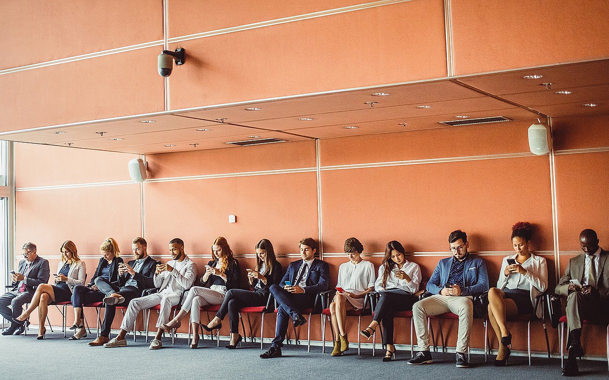 Vor einer orangenen Wand sitzen 13 Personen aufgereiht auf Stühlen und blicken auf ihr Smartphone.