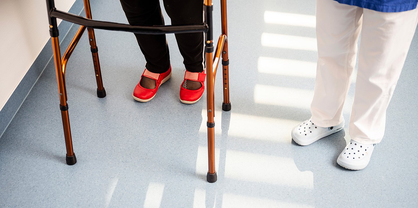 Foto einer Frau mit Gehhilfe und einer Pflegekraft daneben in einem Flur. Man sieht jeweils nur die Beine.