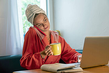 Eine junge Frau sitzt im Bademantel vor dem Laptop, mit Handy am Ohr und einer Kaffeetasse in der Hand. 