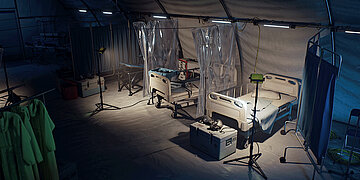In einem gro&szlig;em Zelt sieht man ein Feldlazarett mit mehreren Stationen zur Behandlung von Patienten mit Plast Vorh&auml;ngen und OP Kleidung.