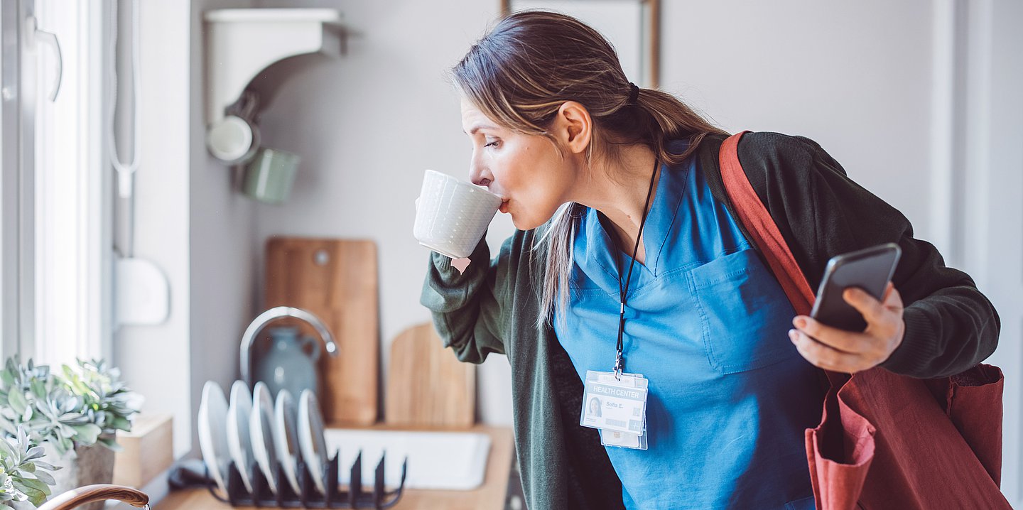 Eine Frau im Mantel und mit umgehängter Tasche trinkt in der Küche stehend noch rasch eine Tasse Kaffee.