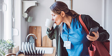 Eine Frau im Mantel und mit umgeh&auml;ngter Tasche trinkt in der K&uuml;che stehend noch rasch eine Tasse Kaffee.