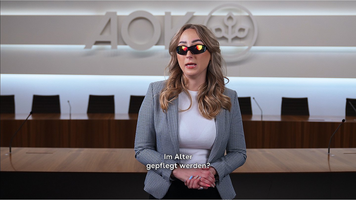 Screenshot: Junge Frau steht im Konferenzsaal des AOK-Bundesverbandes, im Hintergrund das Logo des Verbandes