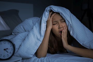 Schlafstörungen in Schleswig-Holstein Foto zeigt eine Frau mitten in der Nacht, die gähnend die Bettdecke über den Kopf gezogen hat und nicht schlafen kann.