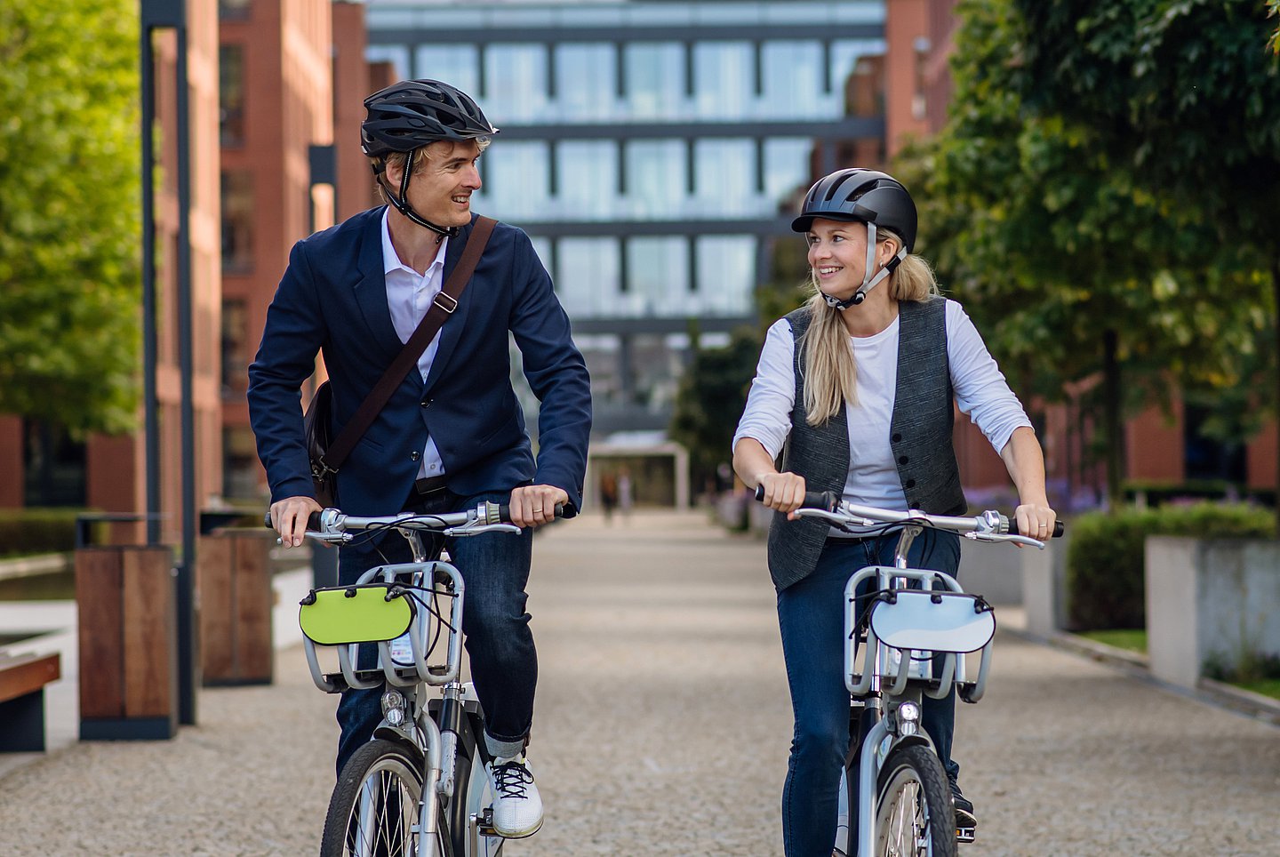 Ein Mann und eine Frau stehen startbereit mit ihren Fahrrädern vor einem Bürogebäude. Beide schauen sich an.Er trägt einen Fahrradhelm, ein dunkeblaues Sakko, darunter ein blassblaues Hemd sowie eine dunkelblaue Jeans und weiße Seaker. Sie trägt ebenfalls einen Fahrradhelm, dazu ein hellgraues Longsleave, eine grau-blau-melierte Weste und blaue Jeans. Im Hintergrund ist in der Unschärfe der Eingang des Gebäudes zu sehen, rechts und links im Anschnitt Bäume. 