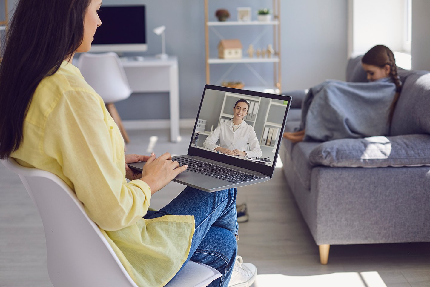 Im Vordergrund sitzt eine Frau/die Mutter auf einem Sessel und spricht per Videosprechstunde mit einem Arzt, der auf dem Bildschirm des Laptops, den sie in der Hand hält, zu sehen ist. Im Hintergrund sitzt auf dem Sofa und in eine Decke eingewickelt, das kranke Kind.