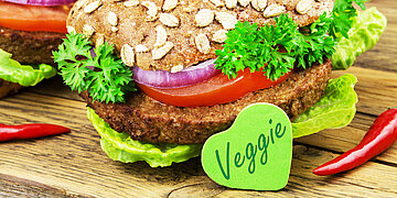 Foto: Auf einem Vollkornbr&ouml;tchen liegt ein vegetarischer Burgerpaddy und Tomaten sowie Salat. Davor steht ein gr&uuml;nes Herz mit der Aufschrift "Veggie".