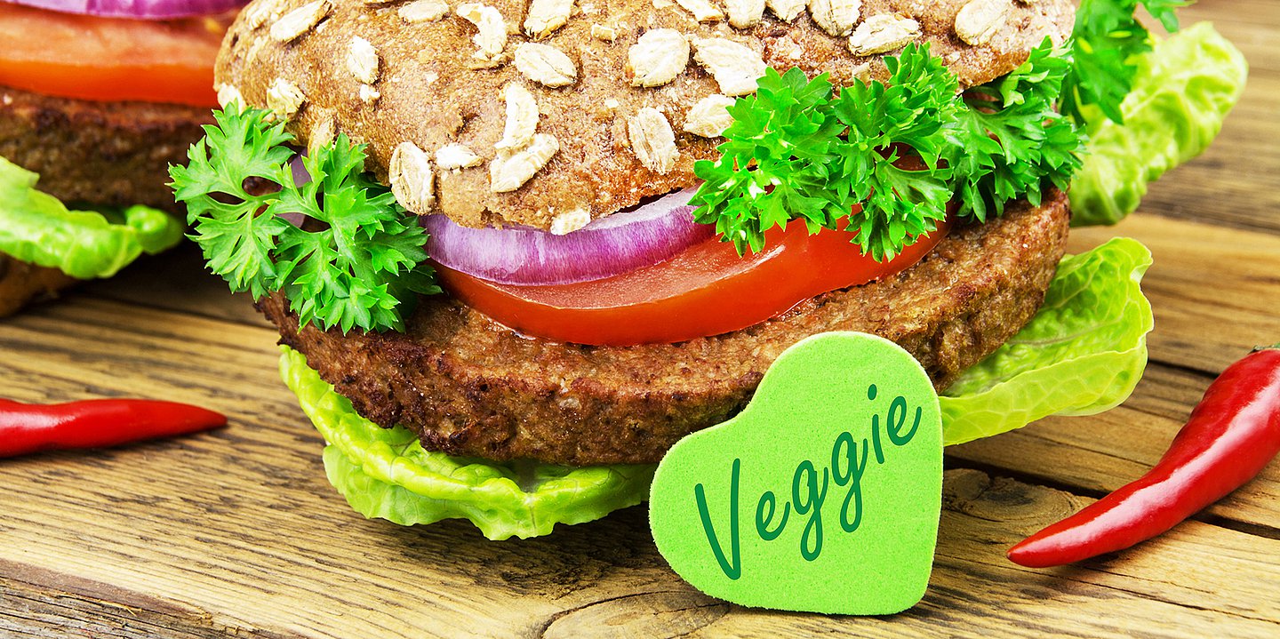 Foto: Auf einem Vollkornbrötchen liegt ein vegetarischer Burgerpaddy und Tomaten sowie Salat. Davor steht ein grünes Herz mit der Aufschrift "Veggie".