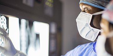 OP-Team bei Röntgen-Diagnose Zwei Ärzte mit OP-Haube und Mundschutz (rechts, Köpfe im Anschniitt) schauen auf ein Rötgenbild, dass an einen Scheinwerfer geklemmt ist.