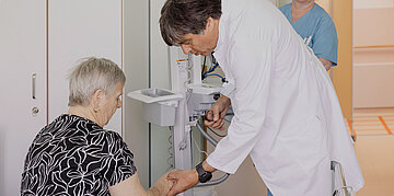 In einem Behandlungszimmer im Krankenhaus sitzt eine &auml;ltere Dame wahrscheinlich auf einem Krankenhausbett oder einem Stuhl. Ein Arzt beugt sich &uuml;ber ihre Hand und f&uuml;hlt ihren Arm.