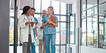 &Uuml;ber einen Krankenhausflur gehen vier Personen: Zwei Frauen vorne (&Auml;rztin und Krankenpflegerin) und zwei M&auml;nner hinten (Arzt und Krankenpfleger). Die &Auml;rztin tr&auml;gt ein Tablet und ein Stethoskop um den Hals und die Krankenpflegerin ein Klemmbrett. Alle tragen die f&uuml;r ihren Beruf typische Kleidung.