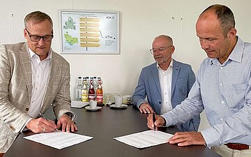 Vertreter der AOK Bodensee-Oberschwaben und des Naturschutzzentrums Wilhelmsdorf unterzeichnen an einem Tisch stehend ihren Kooperationsvertrag.