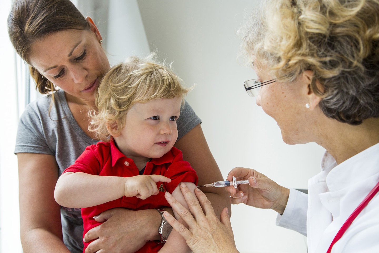 Foto: Ein Kleinkind sitz auf dem Schoß der Mutter und lässt sich von einer Ärztin impfen.