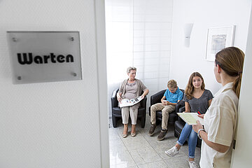 Das Foto zeigt den Blick in ein Wartezimmer mit drei Wartenden und einer MFA. Links h&auml;ngt ein Schild mit der Aufschrift "Warten".