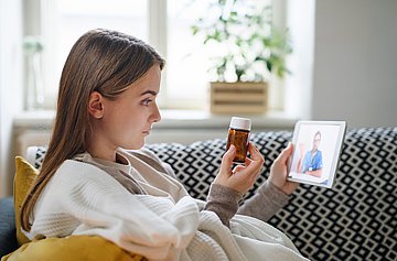 Junge Frau in Videosprechstunde mit ihrem Arzt Junge Frau sitzt zu Hause auf dem Sofa und hat eine Videosprechstunde mit ihrem Arzt. Gerade hält sie ein kleines braunes Medizin-Fläschchen für den Arzt in die Kamera.
