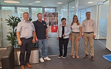 Gruppenbild mit f&uuml;nf stehenden Personen in einem B&uuml;roumfeld.