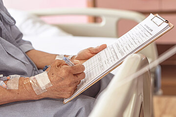 Foto: Ein Patient mit Kan&uuml;len an der rechten Hand f&uuml;llt ein Formular aus. Das Formular ist auf ein Klemmbrett gespannt. 