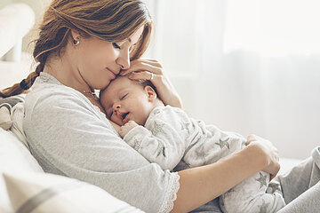 Eine junge Frau liegt im Bett, sie hat ihr Baby auf dem Bauch liegen und h&auml;lt es mit beiden H&auml;nden umfasst. Ihr Kopf ruht am Kopf des Kindes.