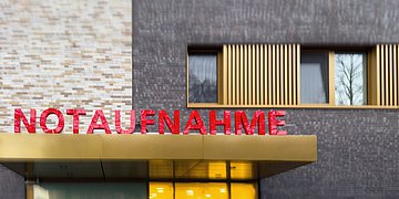 Notaufnahme Der überdachte Eingang eines Krankenhauses: Auf dem Dach prangt in großen, roten Buchstaben das Wort "Notaufnahme". Im Hintergrund ist die Hauswand mit Fenstern zu sehen.