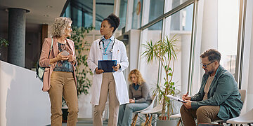 Medizinisches Personal spricht im Wartebereich einer Praxis mit einer Patientin, w&auml;hrend andere Personen im Hintergrund sitzen.