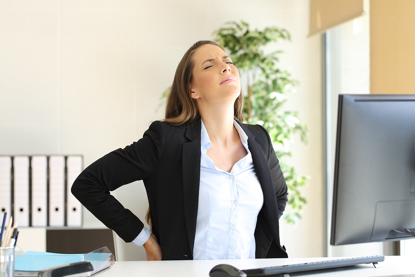 Foto zeigt eine Frau, die im Büro am Schreibtisch sitzt auf dem ein PC steht. Sie hält sich mit beiden Händen den Rücken. Das Gesicht ist leicht nach oben gerichtet und schmerzlich verzogen, die Augen sind geschlossen.