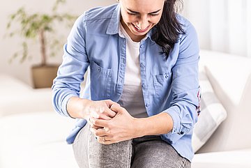 Arthrose in Westfalen-Lippe Eine Frau sitzt mit schmerzverzerrtem Gesicht auf einem Sofa und umfasst mit beiden Händen ihr rechtes, leicht angehobenes Knie.