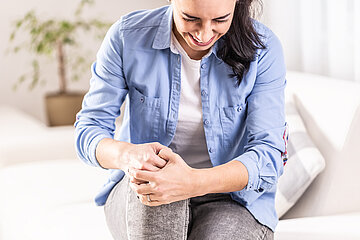Eine Frau sitzt mit schmerzverzerrtem Gesicht auf einem Sofa und umfasst mit beiden H&auml;nden ihr rechtes, leicht angehobenes Knie.