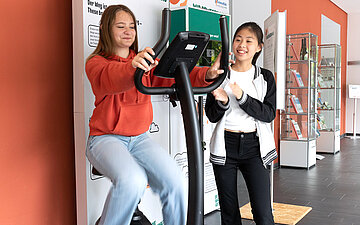 Zwei l&auml;chelnde junge M&auml;dchen probieren ein Fitnessger&auml;t aus.