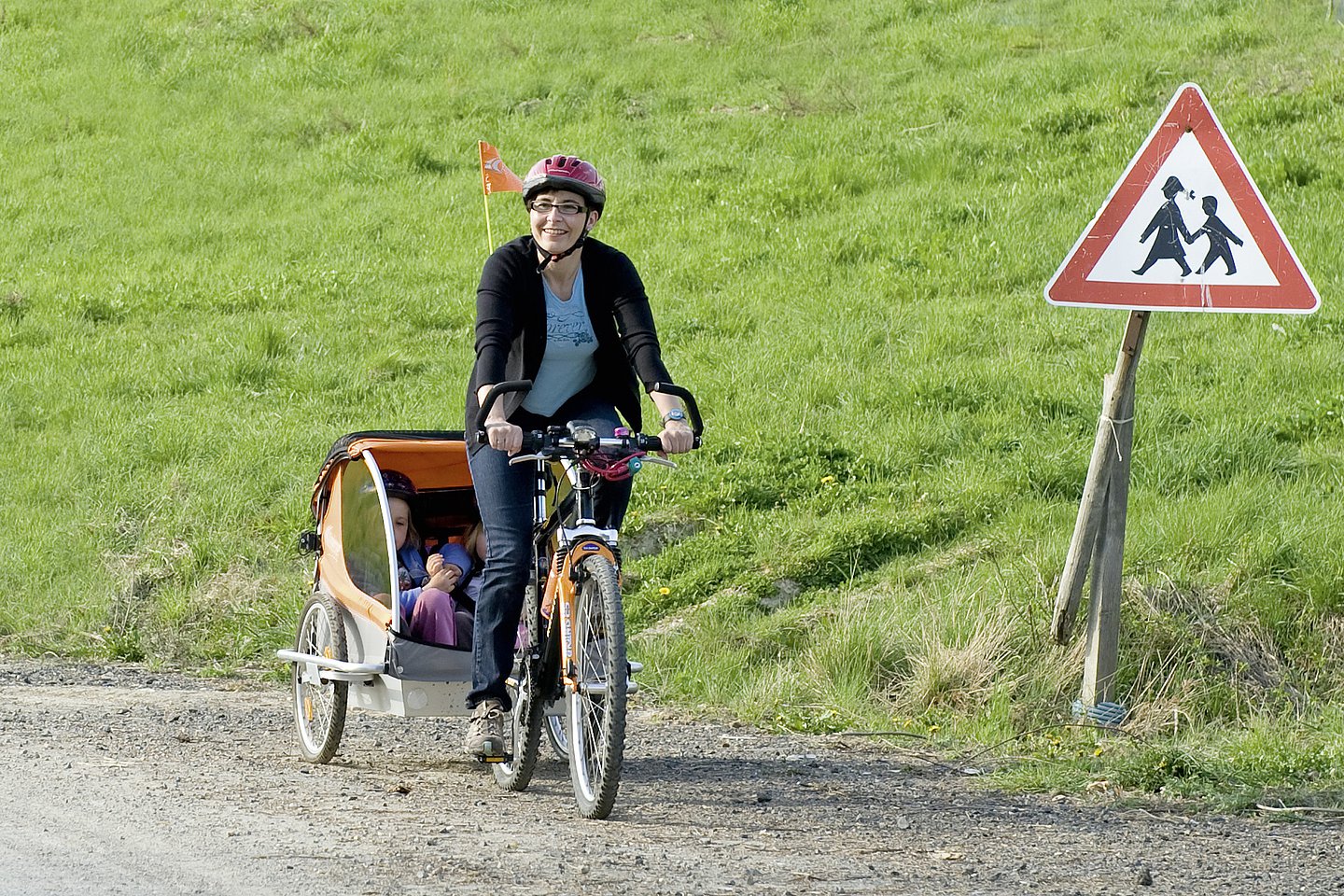 Foto: Eine Frau macht einen Fahrradausflug mit ihren Kindern, die in einem Fahrradanhänger sind. Alle drei Personen tragen einen Schutzhelm.