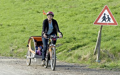 Eine Frau macht einen Fahrradausflug mit ihren Kindern, die in einem Fahrradanhänger sind. Alle drei Personen tragen einen Schutzhelm.