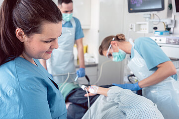 Im Vordergrund ist eine Mitarbeiterin in blauer OP-Kleidung zu sehen, die ihren Blick auf den liegenden Patienten gerichtet hat, bei dem die &Auml;rztin eine Koloskopie durchf&uuml;hrt.