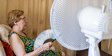 &Auml;ltere Frau sitzt in einem Sessel, f&auml;chelt sich mit einem Handf&auml;cher Luft zu und blickt auf einen gro&szlig;en Ventilator im Vordergrund.