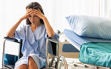 Kopfschmerztag 2025 Schleswig-Holstein Eine Frau, die im Rollstuhl neben ihrem Krankenhausbett sitzt, hält sich die Stirn.