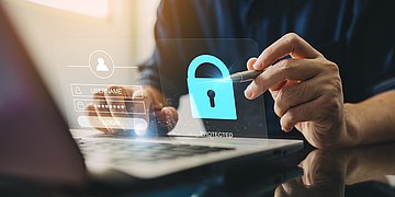 Cyberkriminalität An einem Laptop sitzt eine Person und gibt ihr Passwort ein. In der Bildmitte sind ein Hologramm von der Passworteingabe und ein Schloss zu sehen.