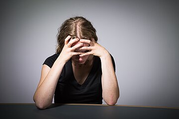 Das Bild zeigt eine junge, blonde Frau mit schwarzem Top an einem Tisch vor einer graus-schattierten Wand sitzend. Sie st&uuml;tzt ihren Kopf auf den Armen und verbirgt dabei ihr Gesicht