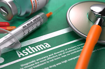 Auf einer Abbildung mit der Aufschrift 'Asthma' liegt ein Stethoskop, eine Spritze und Tabletten in Nahaufnahme. 